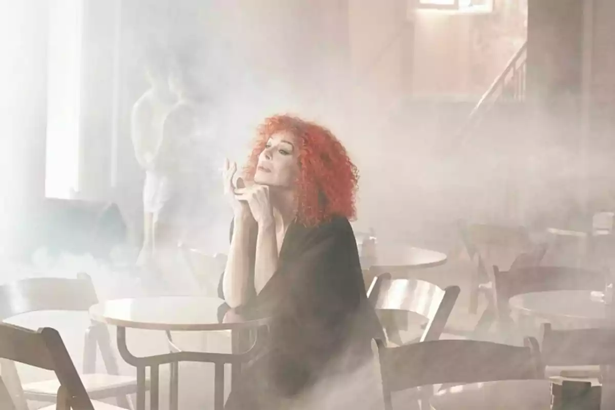 Persona con cabello rizado y rojo sentada en una mesa en un ambiente lleno de humo.