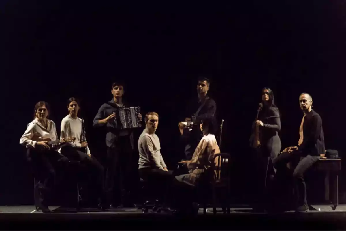 Ocho personas posan en un escenario oscuro, algunas sentadas y otras de pie, varias sostienen instrumentos musicales como guitarra, acordeón y violín, con iluminación tenue que resalta sus figuras. Ocho personas posan en un escenario oscuro, algunas sentadas y otras de pie, varias sostienen instrumentos musicales como guitarra, acordeón y violín, con iluminación tenue que resalta sus figuras.