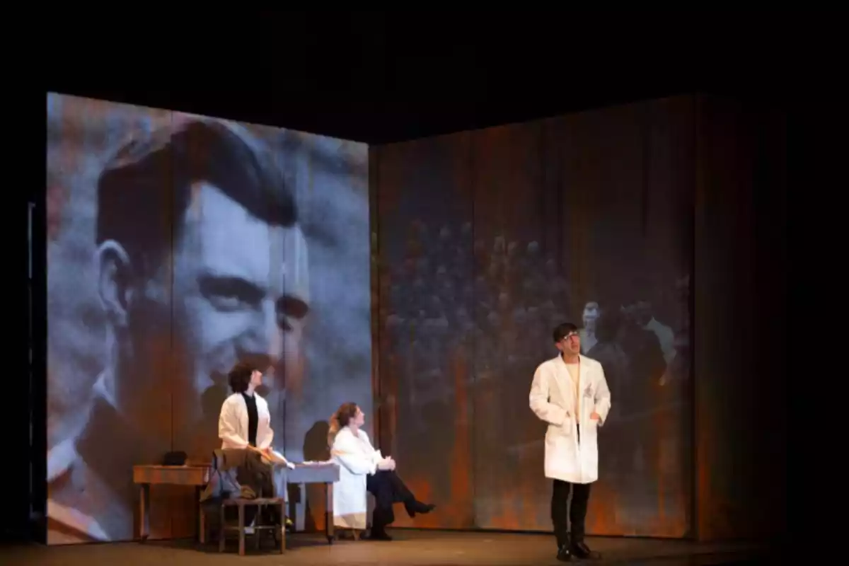 Tres personas con batas blancas actúan en un escenario teatral con proyecciones de imágenes en blanco y negro de fondo, una de un rostro masculino y otra de un grupo de personas