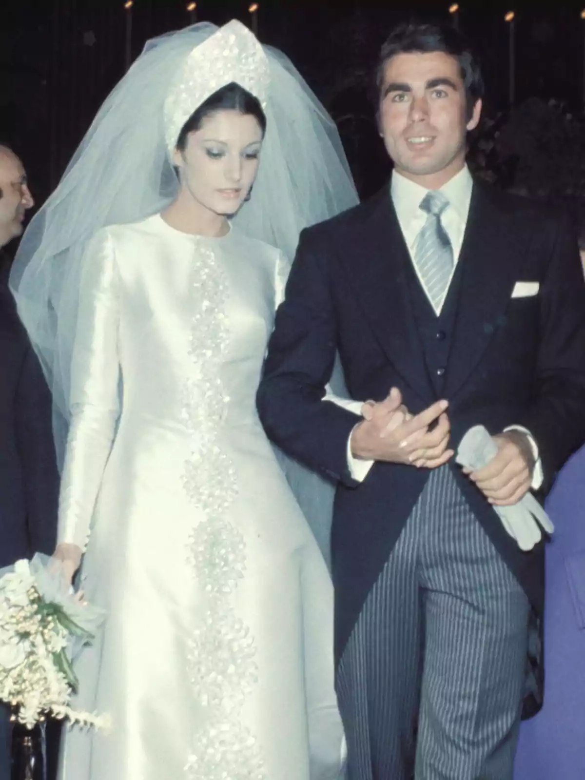 Una pareja vestida de novios, la mujer lleva un vestido blanco con detalles bordados y un velo, mientras que el hombre viste un traje oscuro con corbata.