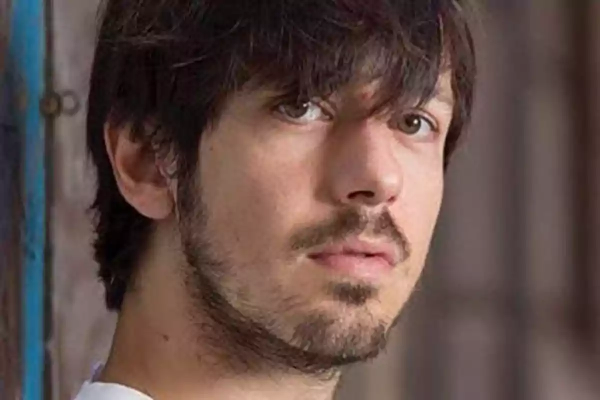 Un hombre joven con barba y cabello oscuro mira hacia la cámara con una expresión seria. Un hombre joven con barba y cabello oscuro mira hacia la cámara con una expresión seria.