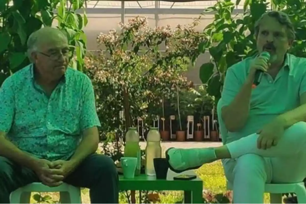 Dos hombres sentados en sillas de plástico, uno de ellos sosteniendo un micrófono, con plantas y vegetación de fondo.