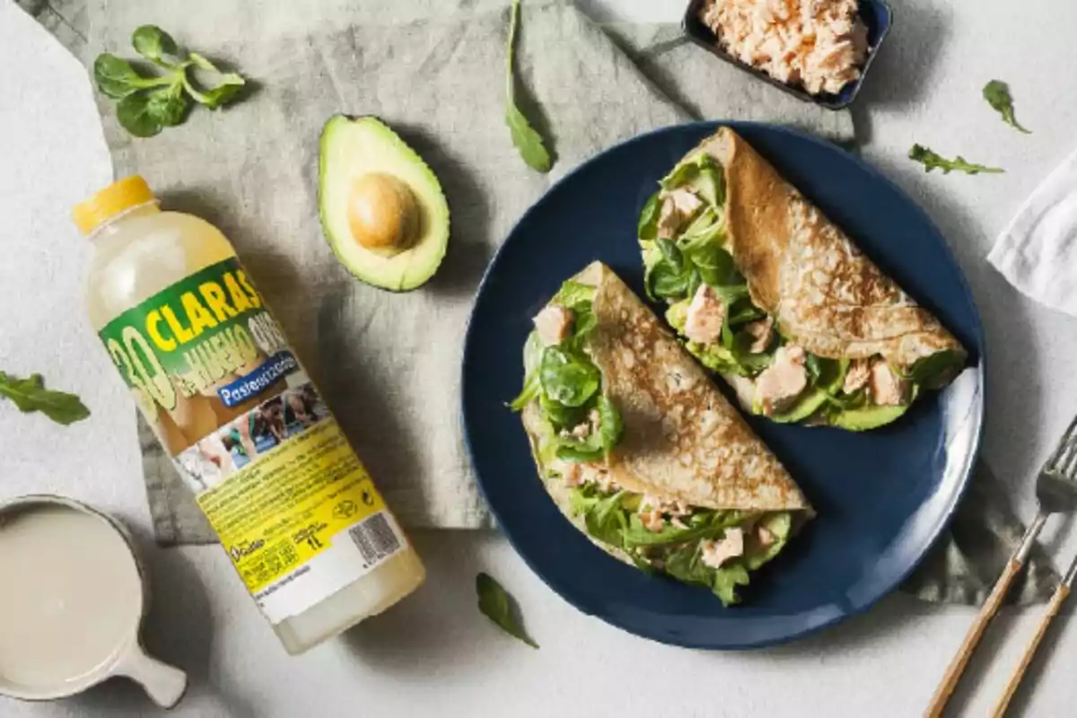 Un plato azul con crepes rellenas de espinacas y salmón junto a un aguacate partido por la mitad una botella de claras de huevo de Mercadona y un recipiente pequeño con más salmón sobre una mesa con un mantel verde. Un plato azul con crepes rellenas de espinacas y salmón junto a un aguacate partido por la mitad una botella de claras de huevo de Mercadona y un recipiente pequeño con más salmón sobre una mesa con un mantel verde.