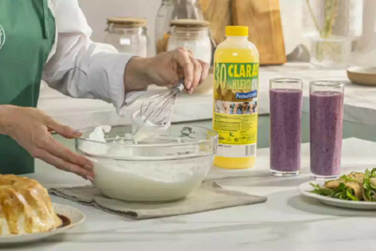 Una persona con delantal verde está batiendo claras de huevo en un tazón de vidrio en una cocina, junto a una botella de claras de huevo y dos vasos de batido morado. Una persona con delantal verde está batiendo claras de huevo en un tazón de vidrio en una cocina, junto a una botella de claras de huevo y dos vasos de batido morado.