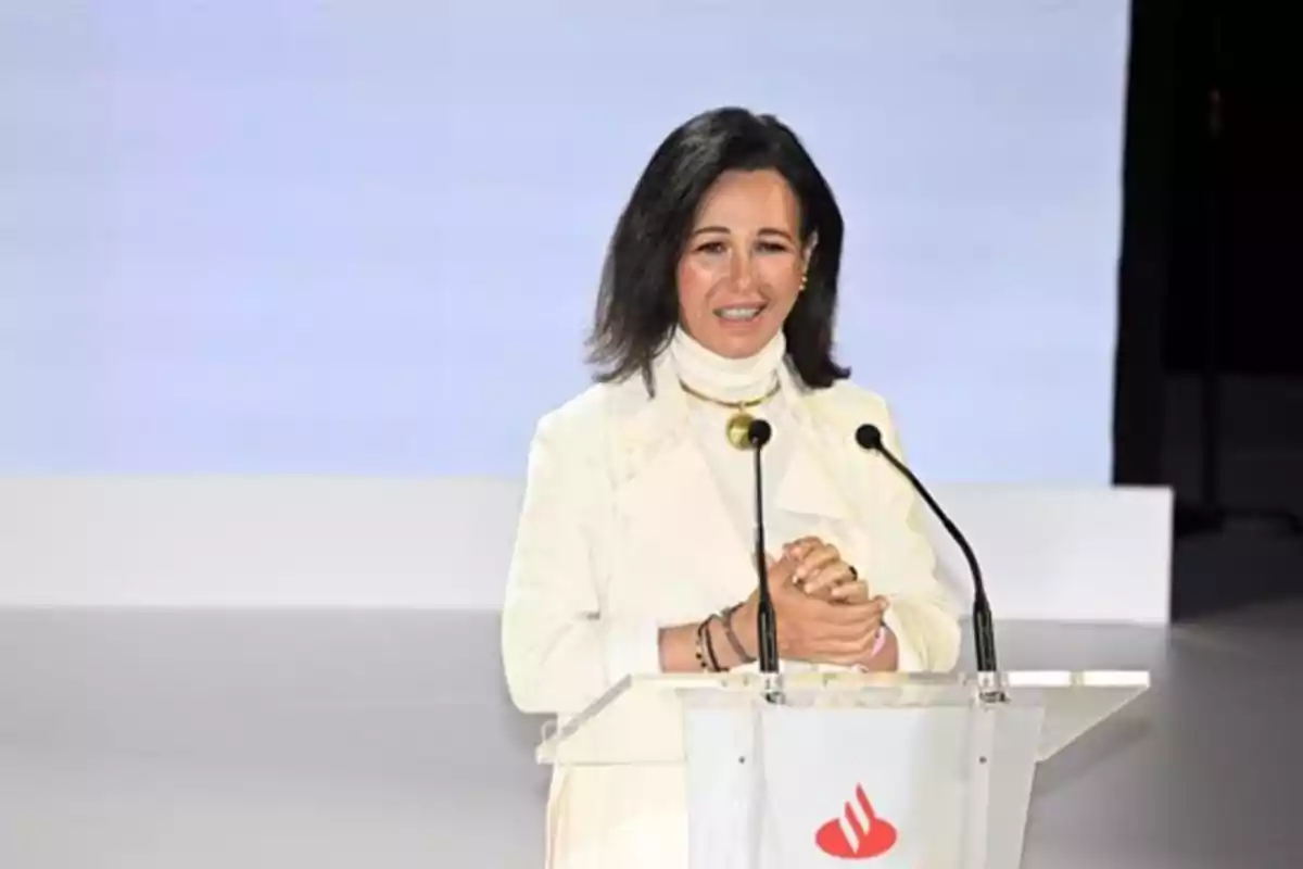 Una persona de pie detrás de un podio con micrófonos, vestida de blanco y sonriendo.
