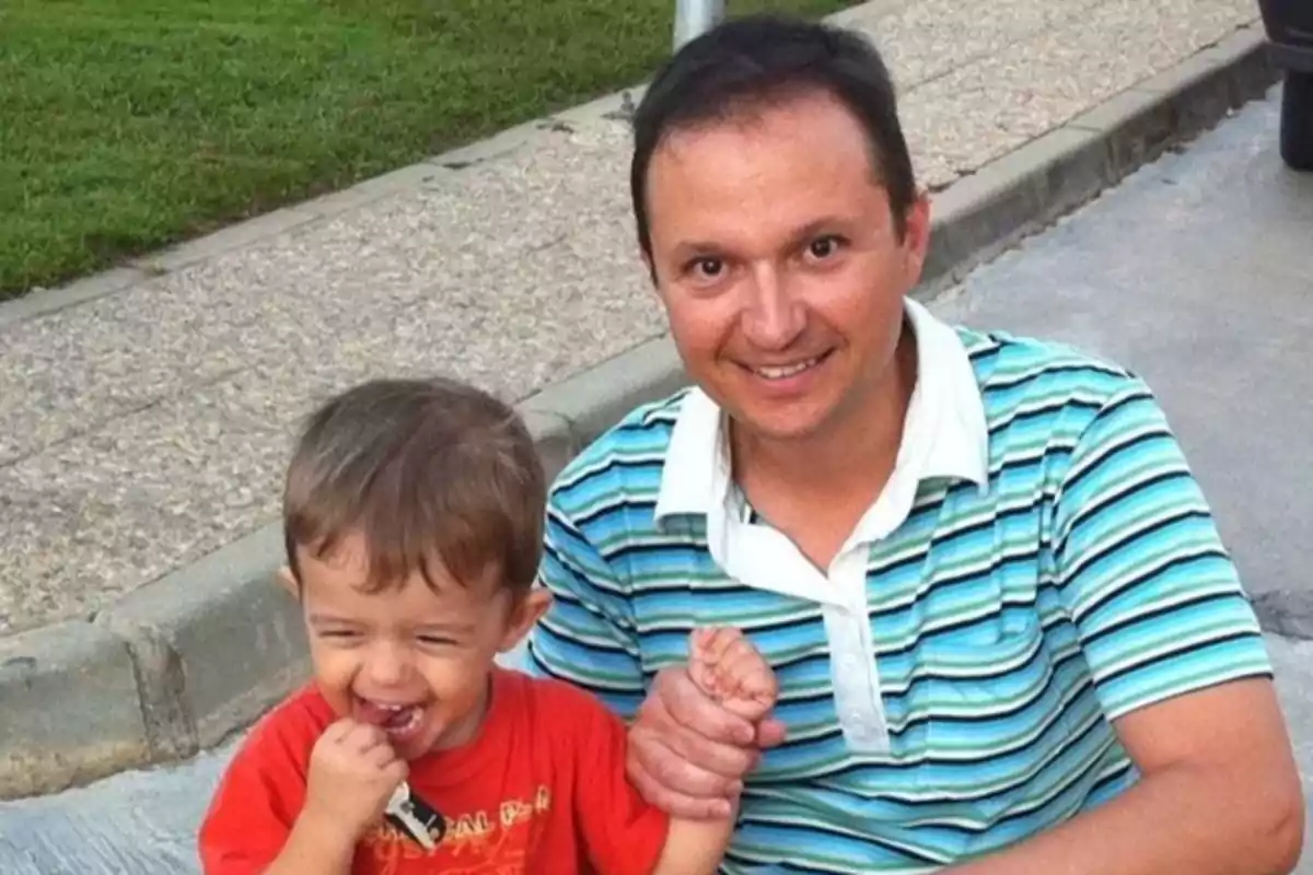 Un hombre sonriente con una camiseta de rayas sostiene la mano de un niño pequeño que lleva una camiseta roja y está riendo mientras muerde un juguete.