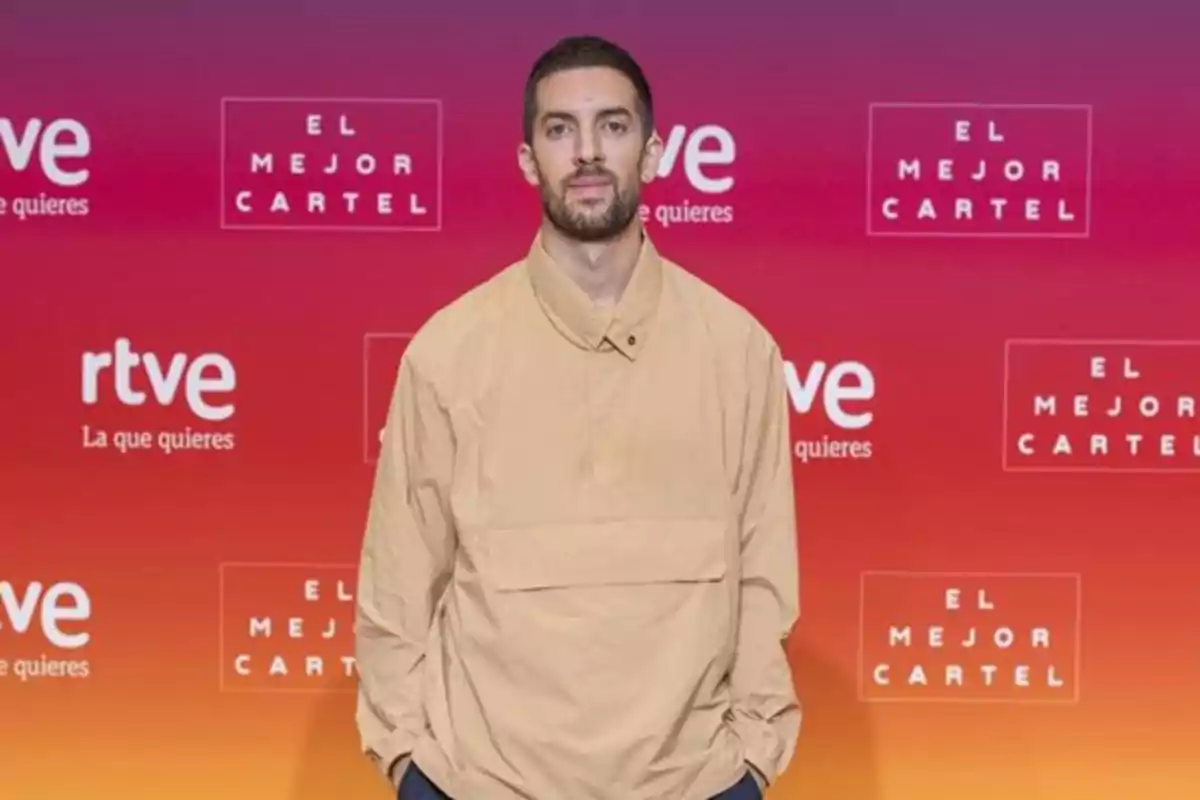 Un hombre con barba y cabello corto lleva una chaqueta beige y posa frente a un fondo con logotipos de RTVE y 