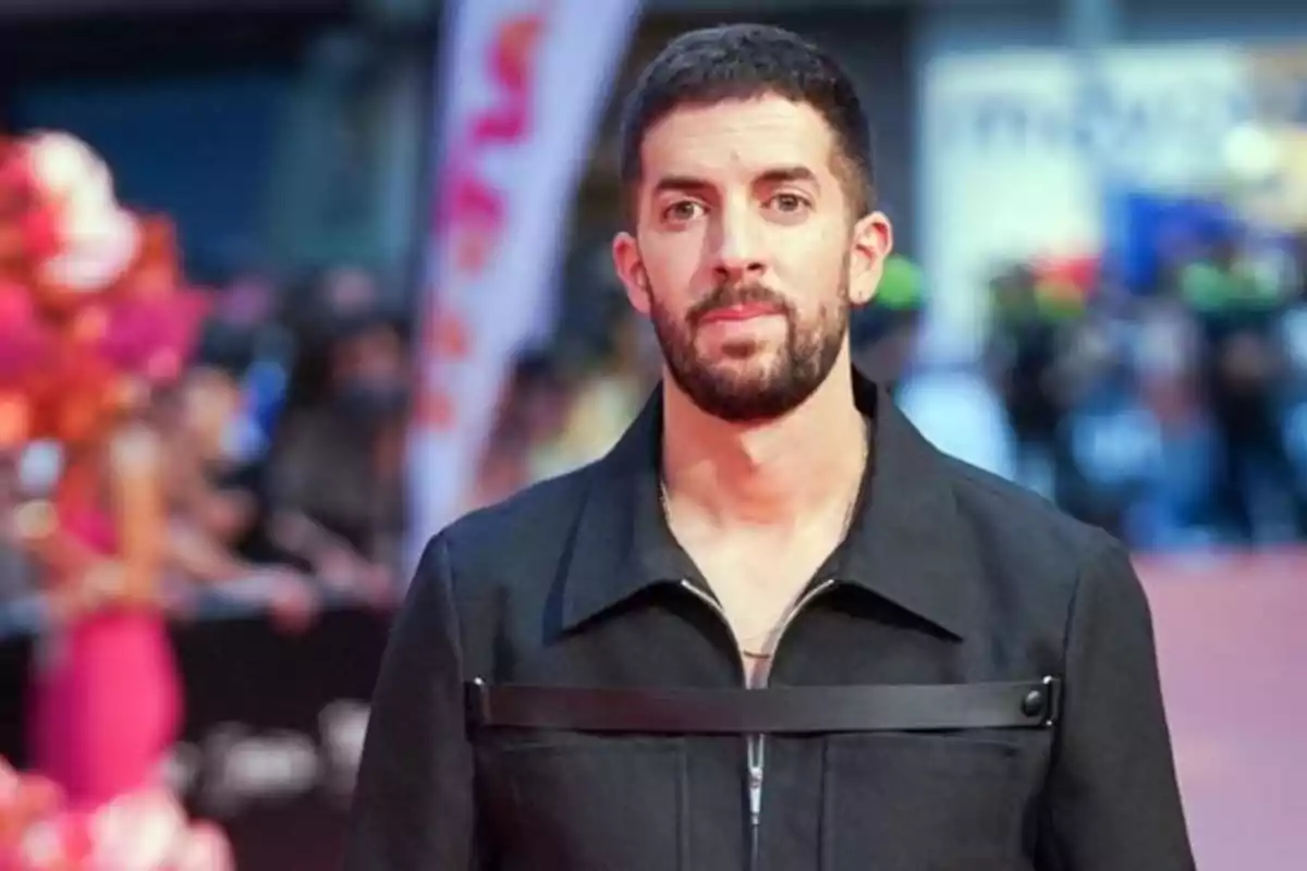 Un hombre con barba y cabello corto viste una chaqueta oscura mientras posa en una alfombra roja con un fondo desenfocado.