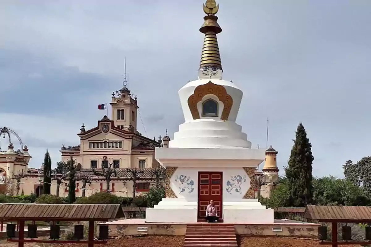 Una persona meditando frente a una estupa blanca con detalles dorados y un edificio histórico al fondo.