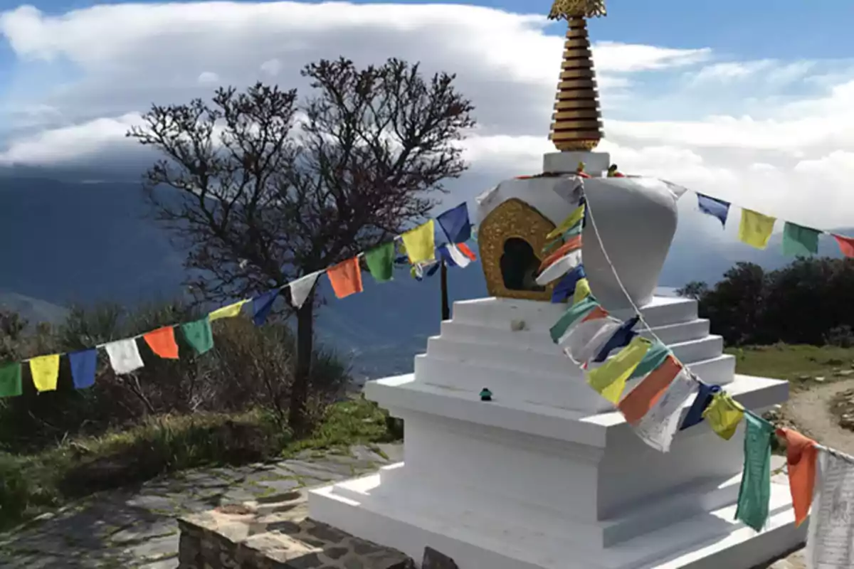 Una estupa blanca con banderas de oración de colores en un entorno montañoso con cielo parcialmente nublado.