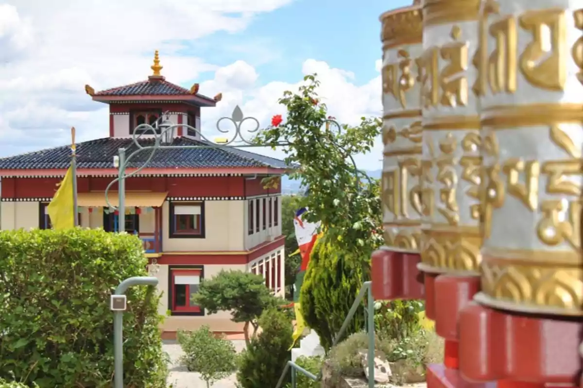 Un templo budista con ruedas de oración doradas en primer plano y un edificio tradicional con techo azul y detalles rojos en el fondo.