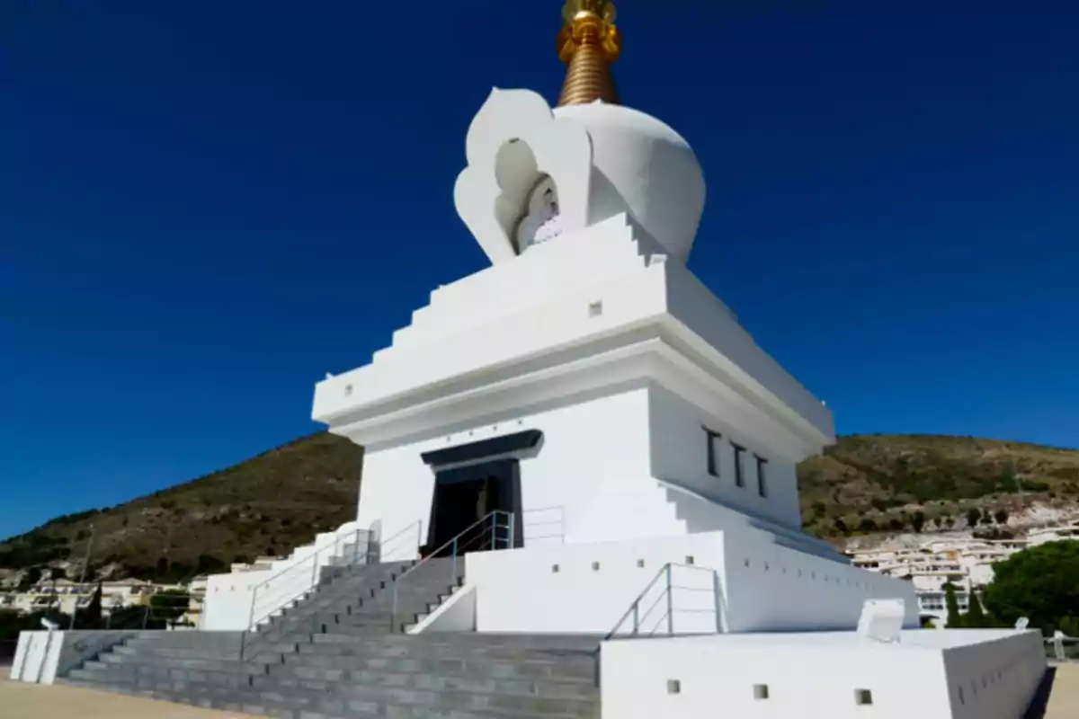 Una estupa blanca con una estructura dorada en la cima, situada en un entorno montañoso bajo un cielo despejado.