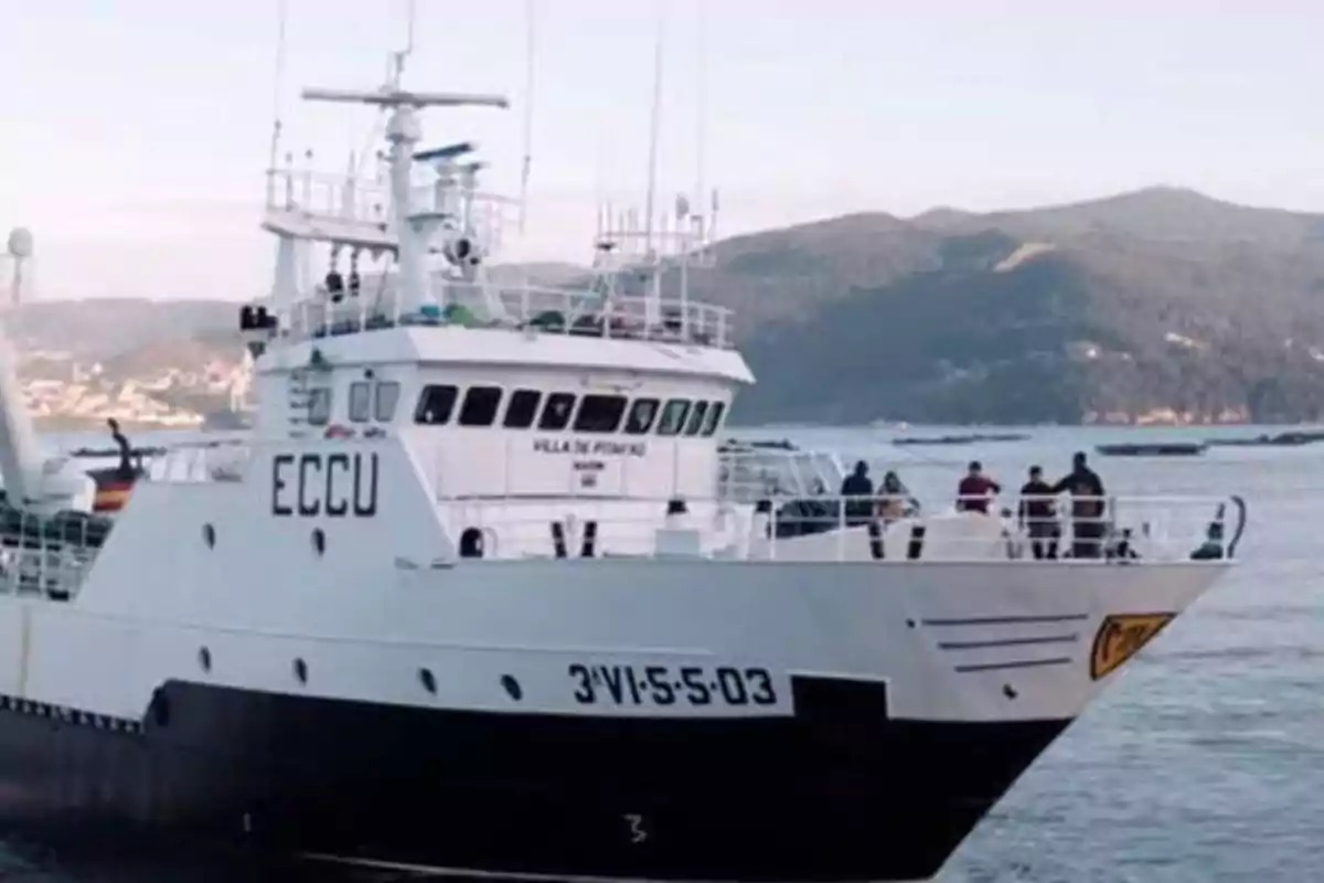 Un barco pesquero blanco y negro con la inscripción "ECCU" en el costado, navegando en un cuerpo de agua con montañas al fondo.