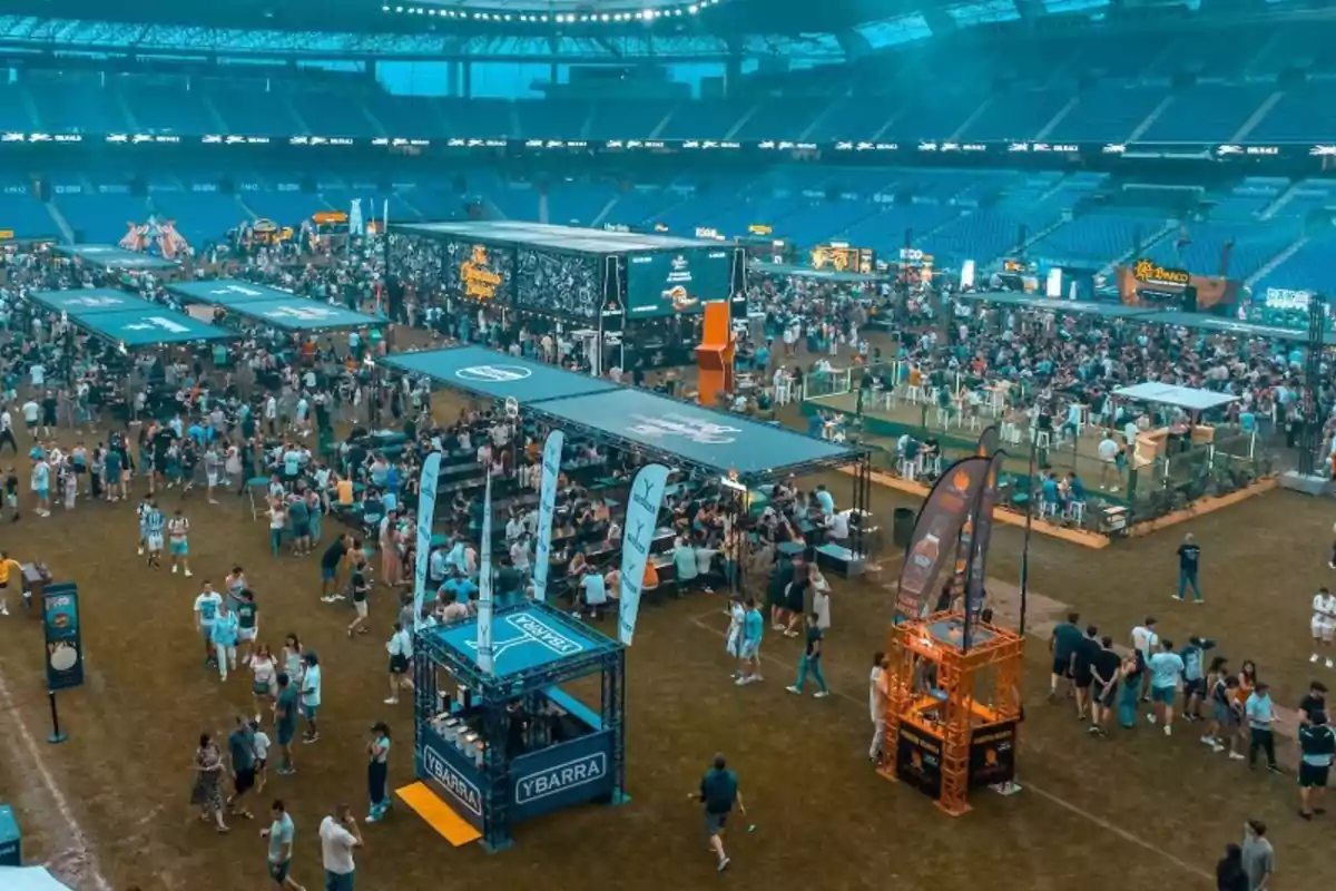 Una multitud de personas disfruta de un evento en un estadio con puestos y carpas distribuidos en el campo.