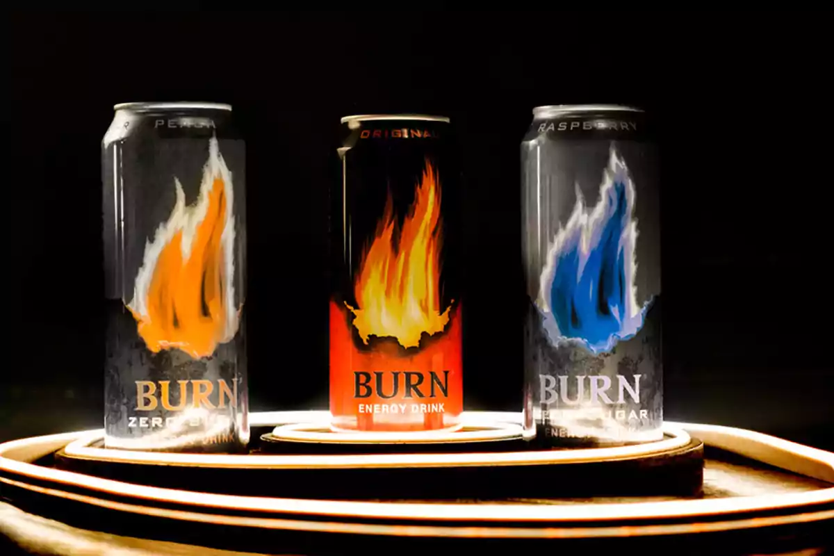 Tres latas de bebida energética Burn con diferentes sabores y colores de llamas en un fondo oscuro.