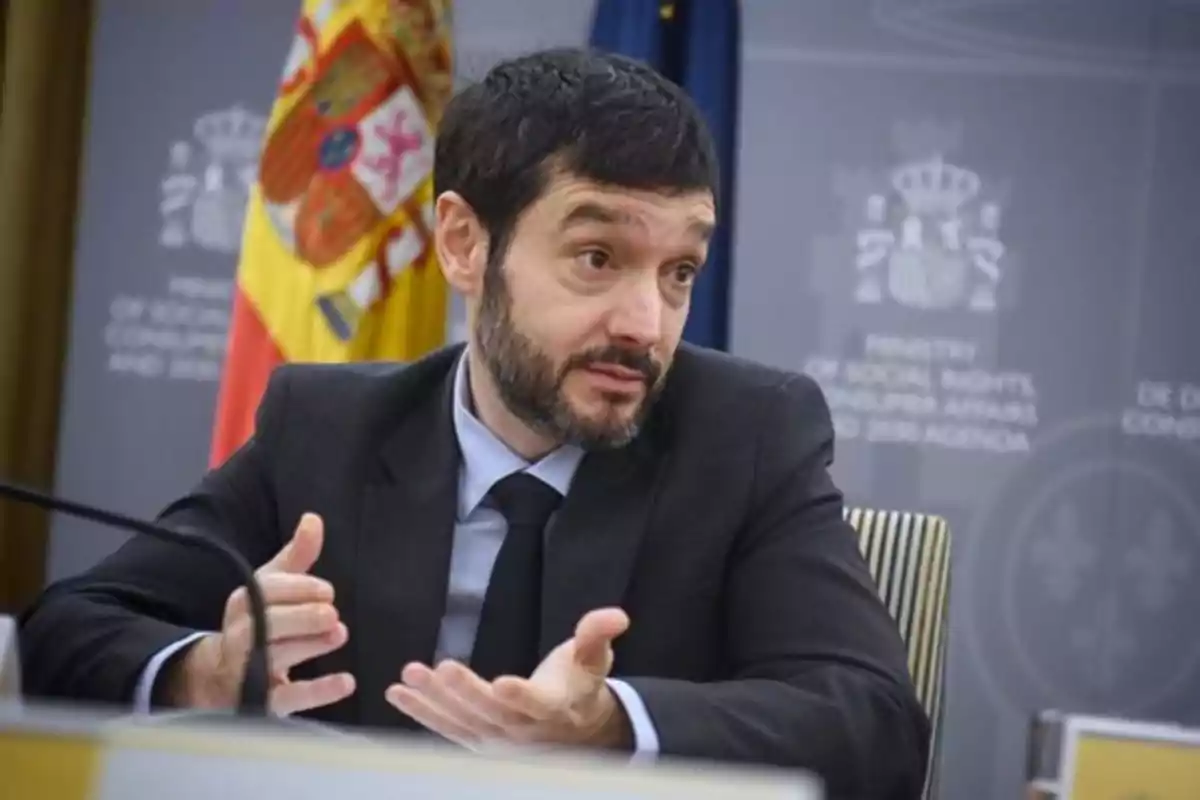 Hombre con barba y traje oscuro hablando en una rueda de prensa con una bandera de España al fondo Hombre con barba y traje oscuro hablando en una rueda de prensa con una bandera de España al fondo