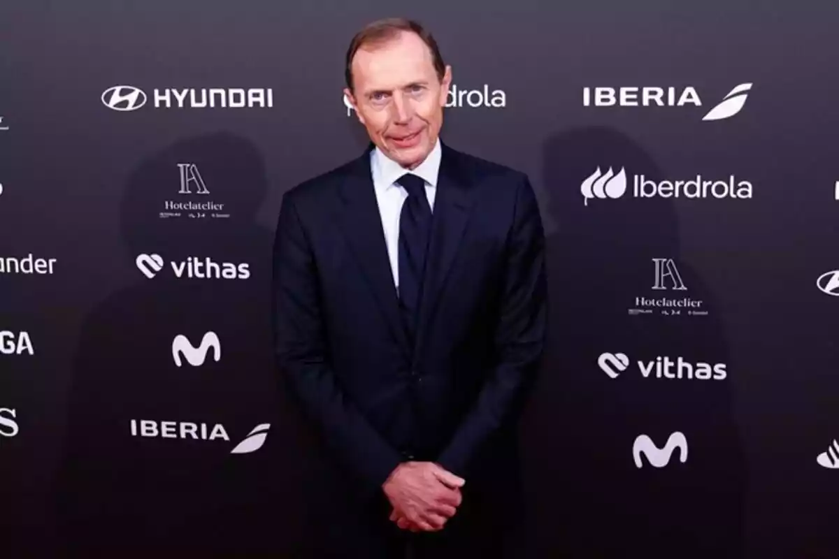 Un hombre con traje oscuro posa frente a un fondo con logotipos de varias marcas. Un hombre con traje oscuro posa frente a un fondo con logotipos de varias marcas.