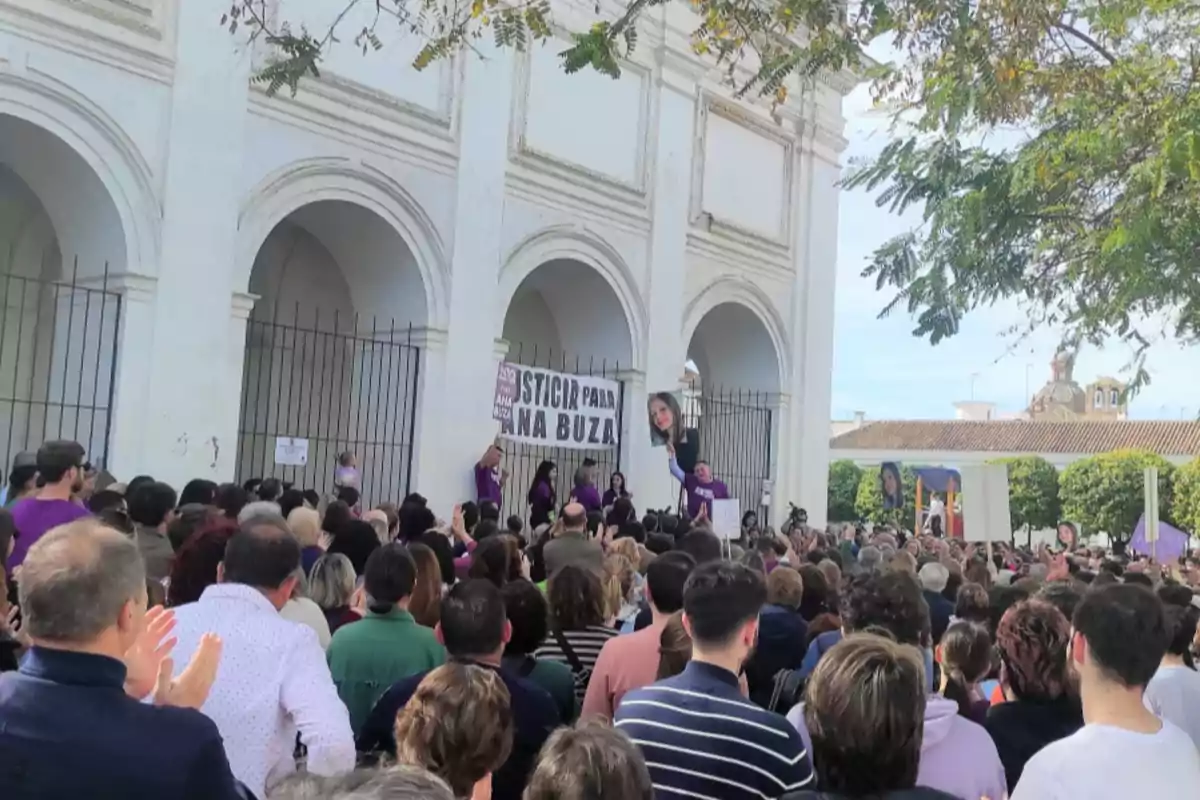 Una multitud se reúne frente a un edificio con arcos sosteniendo pancartas y carteles en una manifestación.