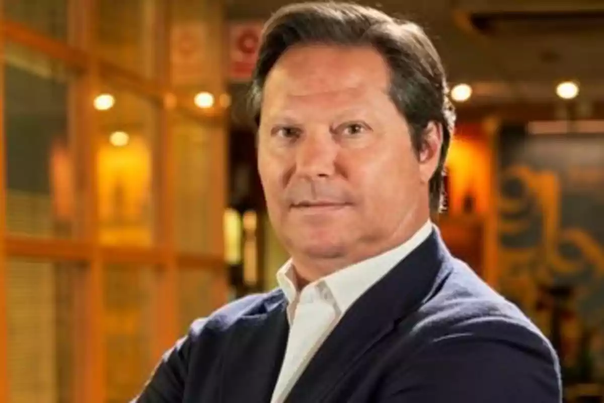 Hombre de mediana edad con cabello oscuro y peinado hacia atrás, vestido con saco azul y camisa blanca, posando con los brazos cruzados en un entorno interior iluminado. Hombre de mediana edad con cabello oscuro y peinado hacia atrás, vestido con saco azul y camisa blanca, posando con los brazos cruzados en un entorno interior iluminado.