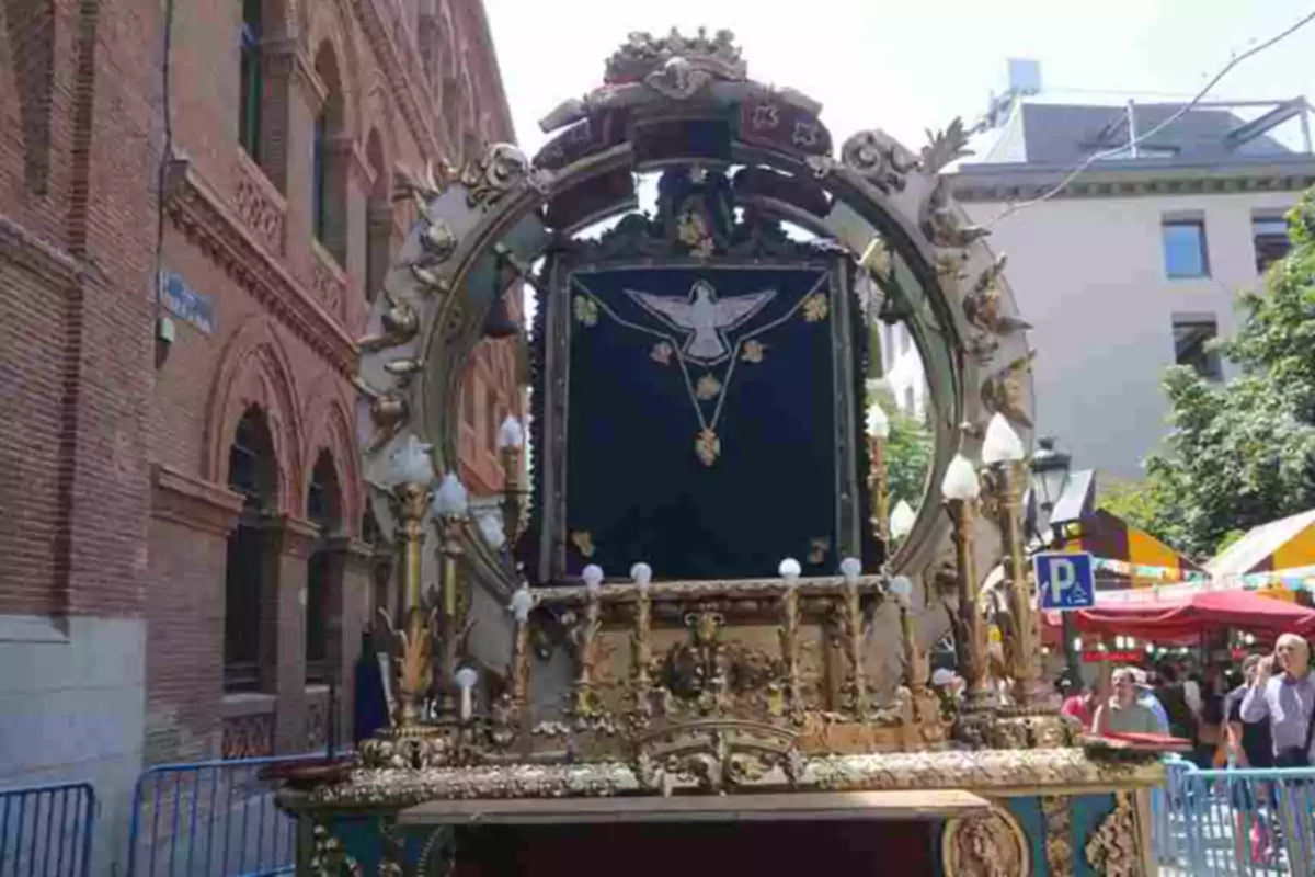 Carroza religiosa adornada con detalles dorados y una imagen de una paloma en el centro, en una calle con edificios de ladrillo y personas alrededor.