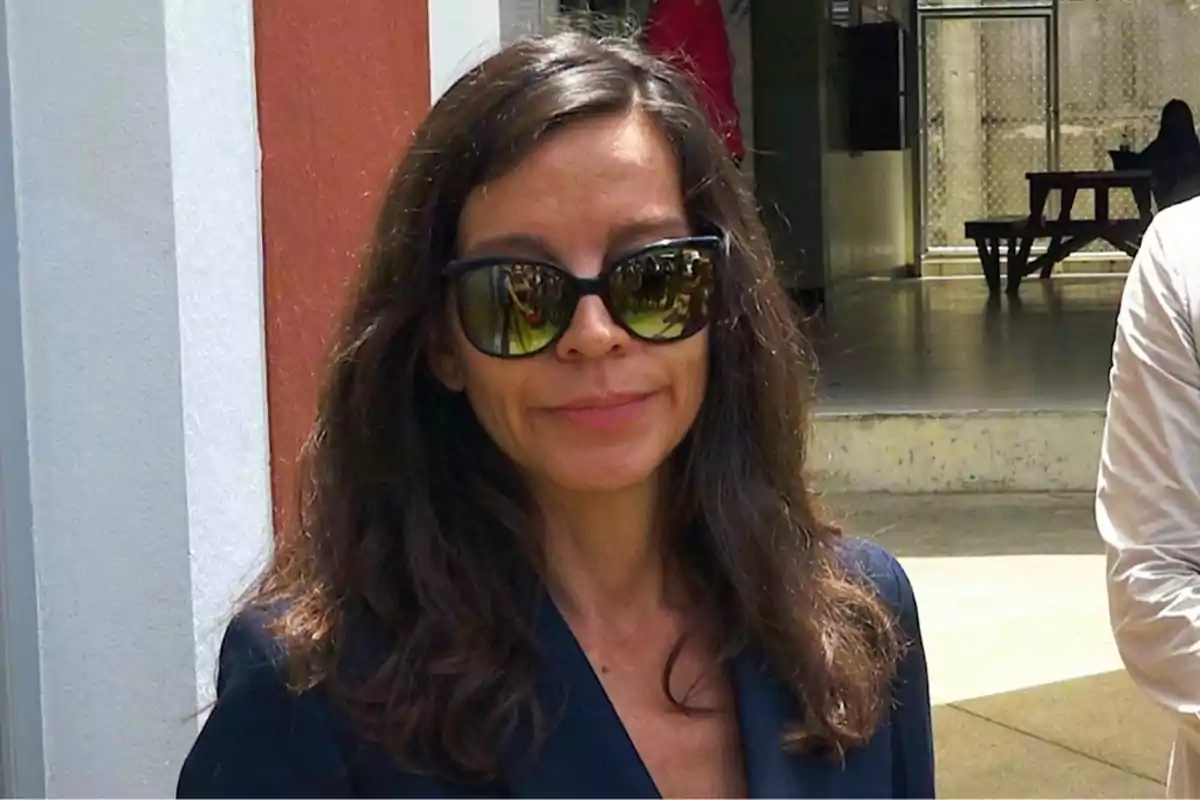 Una mujer con gafas de sol oscuras y cabello largo y suelto está de pie frente a un edificio con una pared de color rojo y blanco.