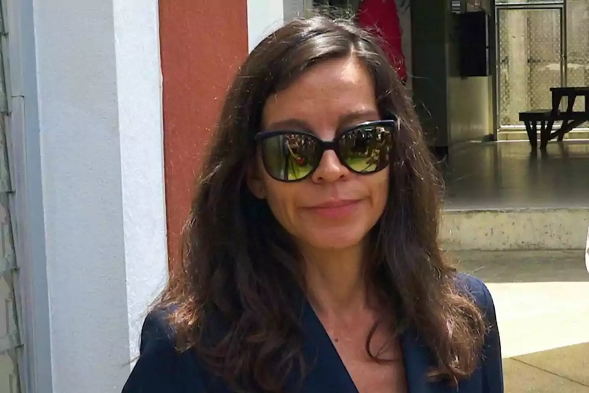 Mujer con gafas de sol y cabello largo y oscuro, de pie frente a un edificio con paredes blancas y rojas.