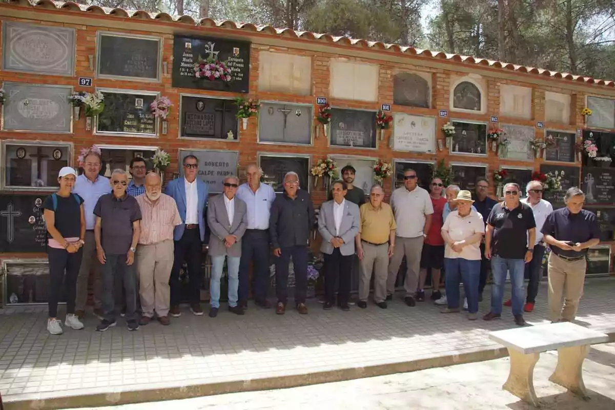 Un grupo de personas posa frente a un muro de nichos en un cementerio, algunos llevan gafas de sol y ropa casual, mientras que otros visten de manera más formal, y hay flores decorando varias lápidas. Un grupo de personas posa frente a un muro de nichos en un cementerio, algunos llevan gafas de sol y ropa casual, mientras que otros visten de manera más formal, y hay flores decorando varias lápidas.