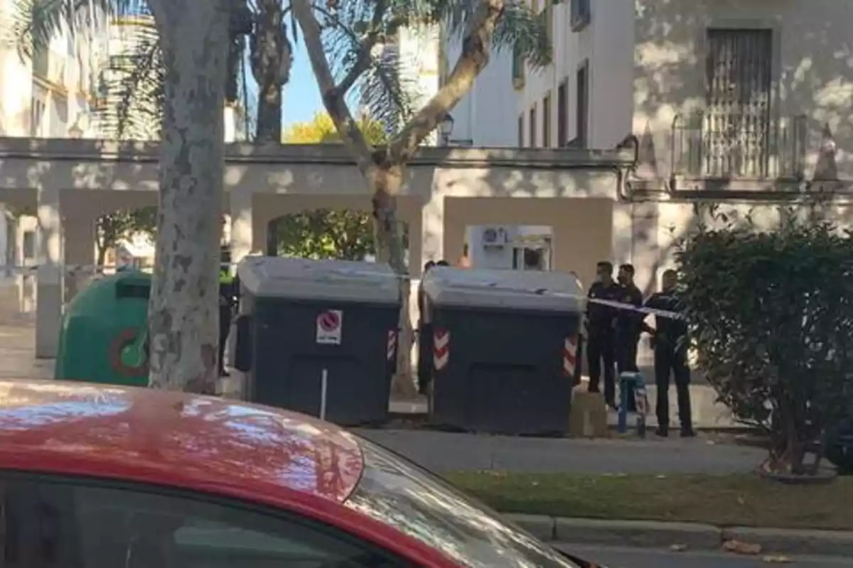 Una escena urbana con contenedores de basura, un coche rojo en primer plano y varios policías al fondo.