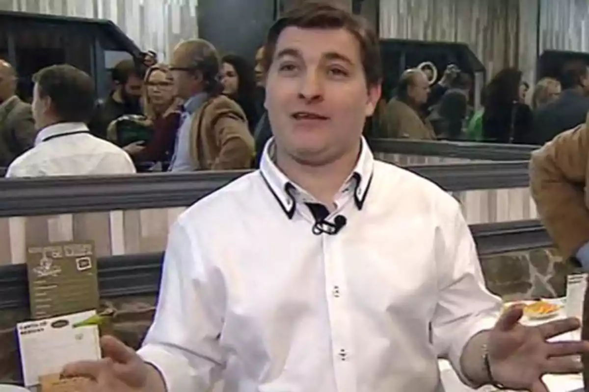 Un hombre con camisa blanca y micrófono de solapa está hablando en un lugar concurrido con varias personas al fondo y un espejo grande reflejando la escena. Un hombre con camisa blanca y micrófono de solapa está hablando en un lugar concurrido con varias personas al fondo y un espejo grande reflejando la escena.
