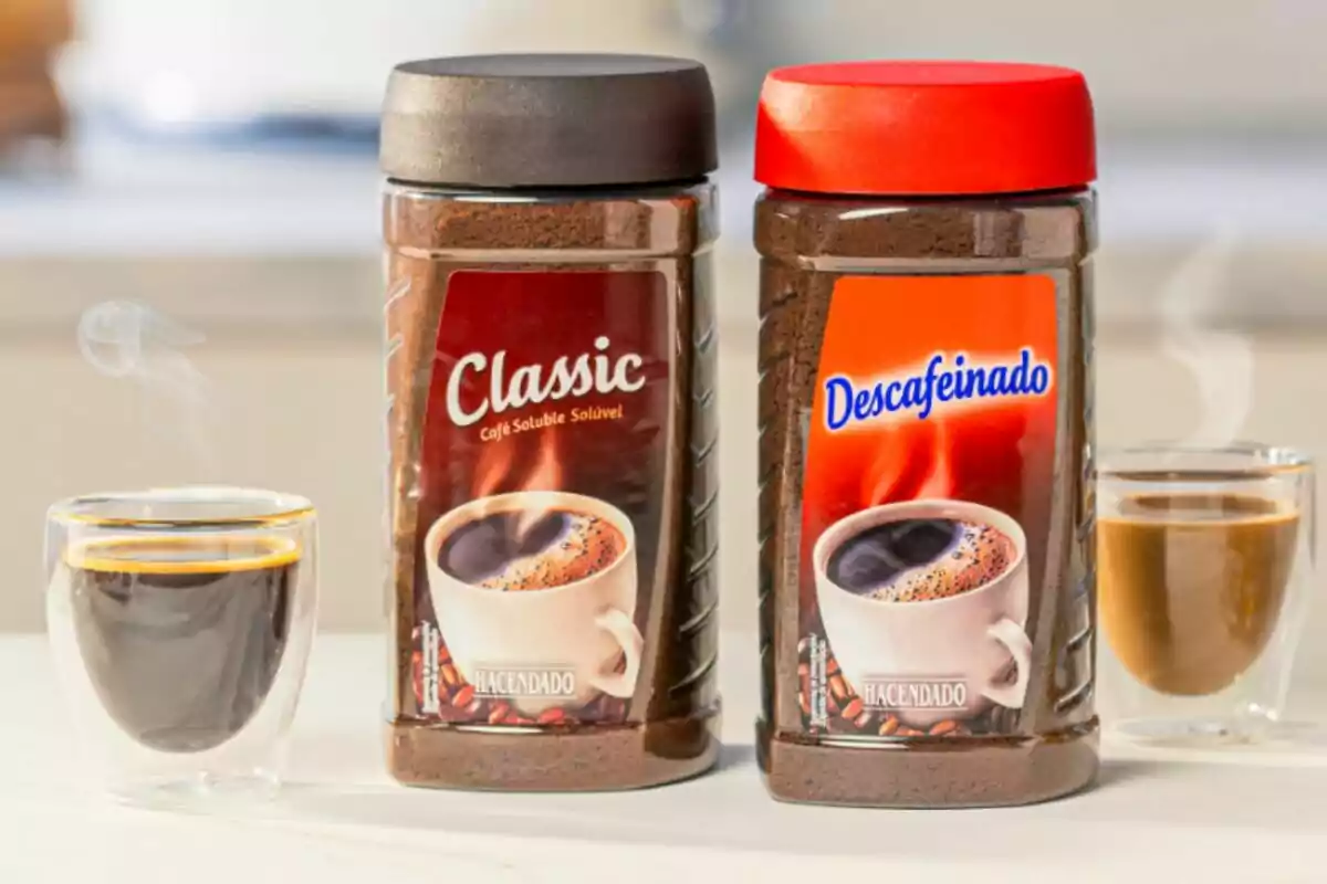 Dos frascos de café soluble, uno clásico y otro descafeinado, con tazas de café humeante al frente. Dos frascos de café soluble, uno clásico y otro descafeinado, con tazas de café humeante al frente.
