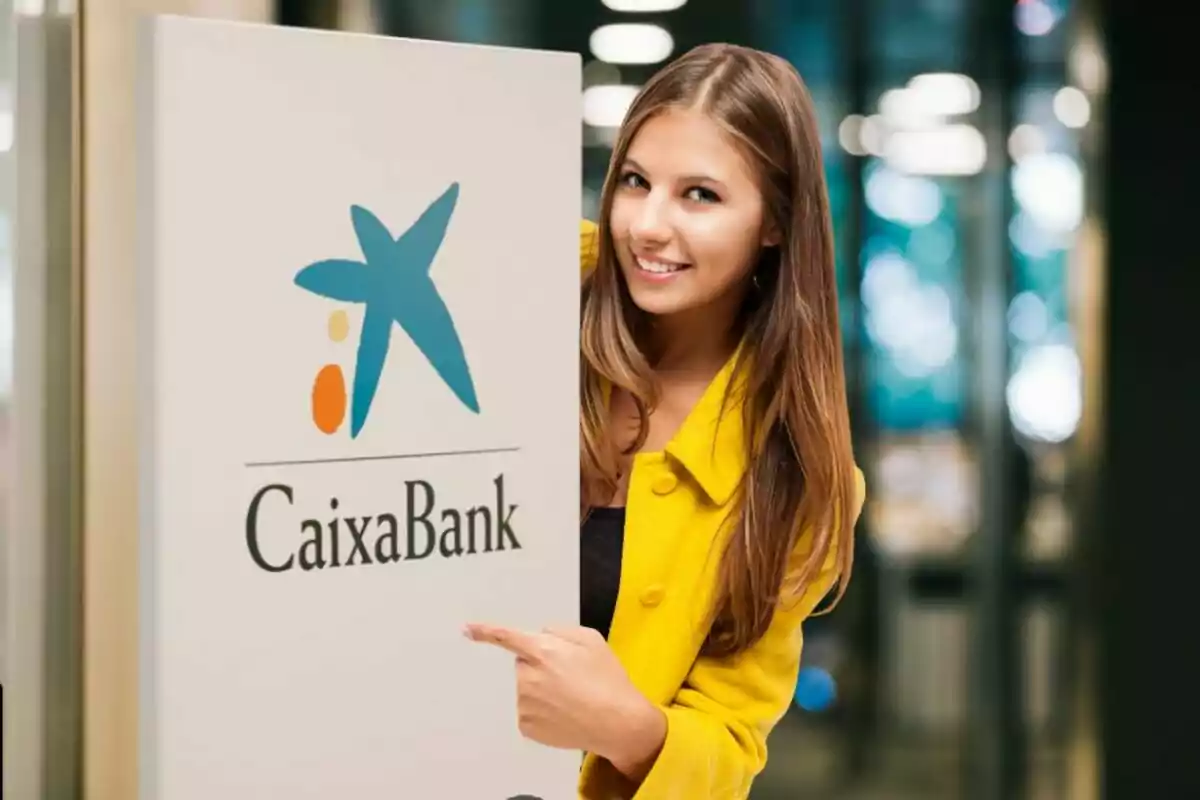 Una mujer con una chaqueta amarilla sonríe y señala un cartel de CaixaBank.