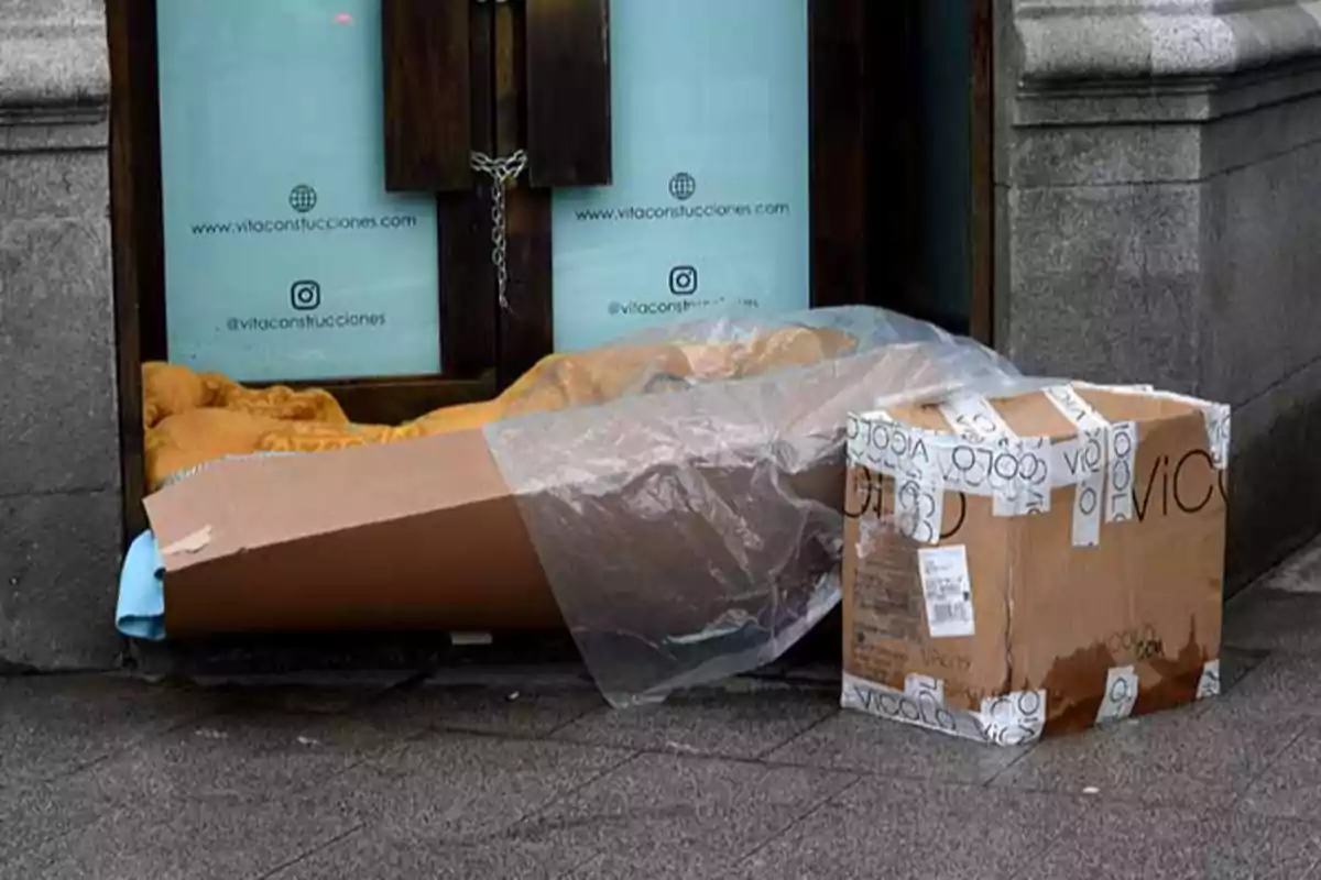 Una caja de cartón y un plástico cubren un espacio en la entrada de un edificio con ventanas de vidrio y paredes de piedra.