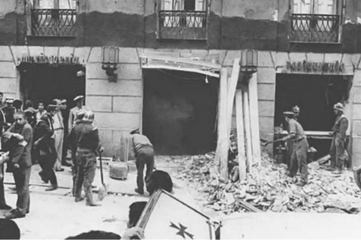 Un grupo de personas, incluyendo soldados y civiles, se encuentra frente a un edificio dañado, con escombros en el suelo y algunos individuos trabajando en la limpieza y reparación del lugar.