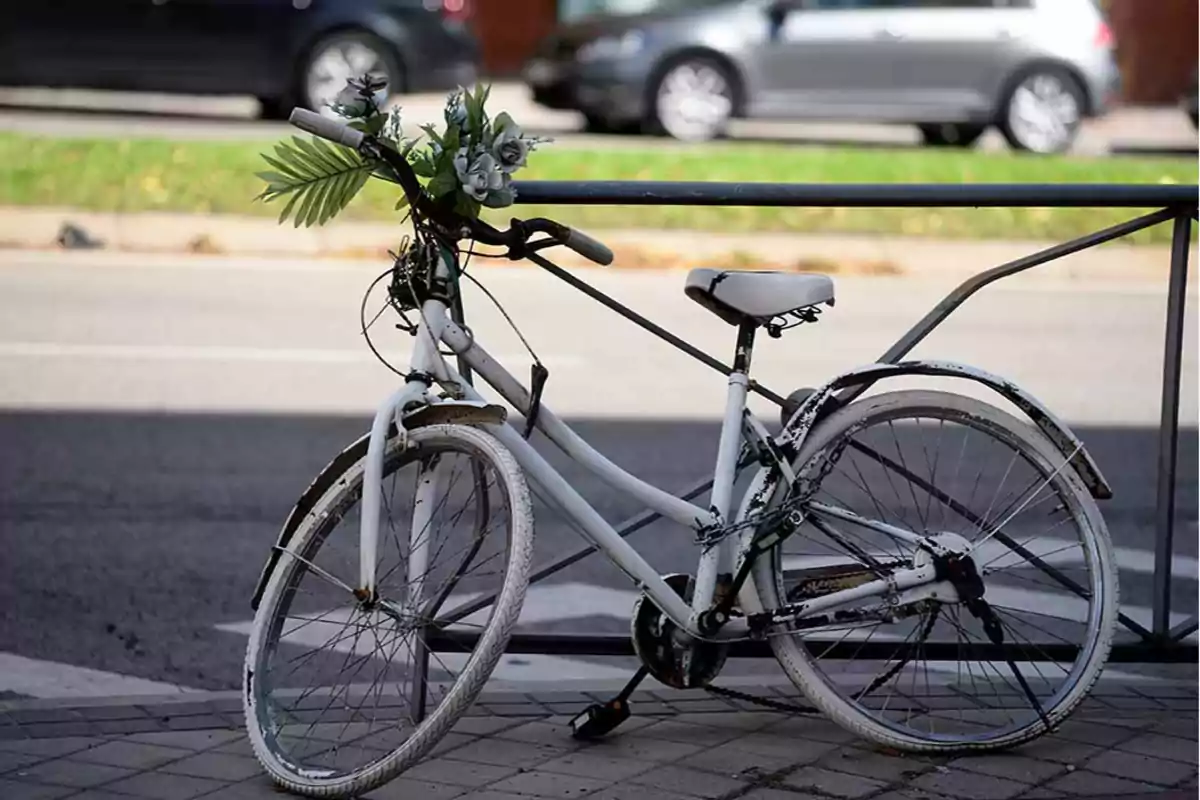 Una bicicleta blanca decorada con flores está asegurada a una barandilla junto a una calle.