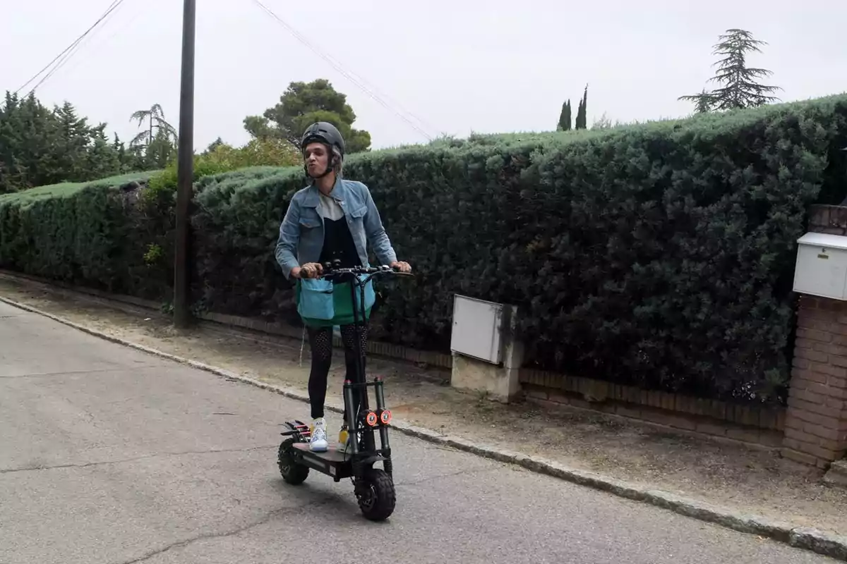 Una persona con casco monta un scooter eléctrico en una calle residencial con setos altos y árboles al fondo.
