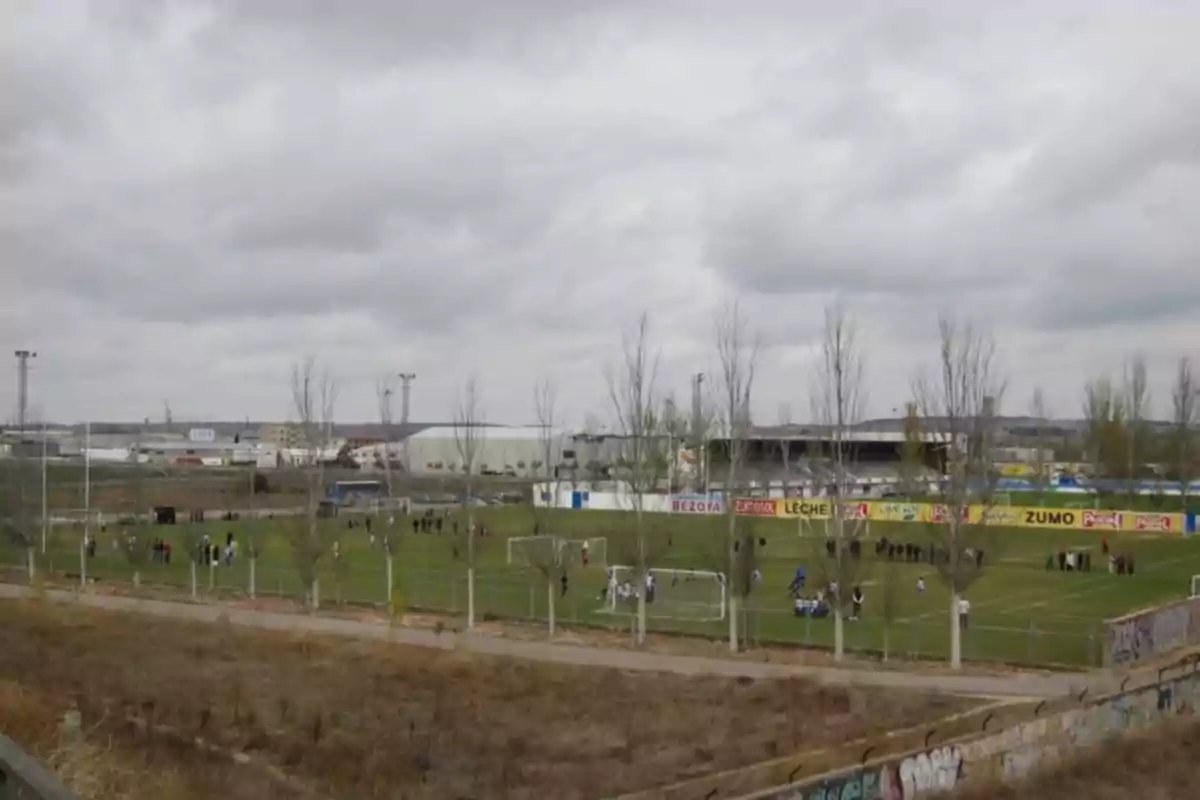 Un campo de fútbol con varias personas jugando y entrenando rodeado de una cerca y con edificios industriales al fondo bajo un cielo nublado.