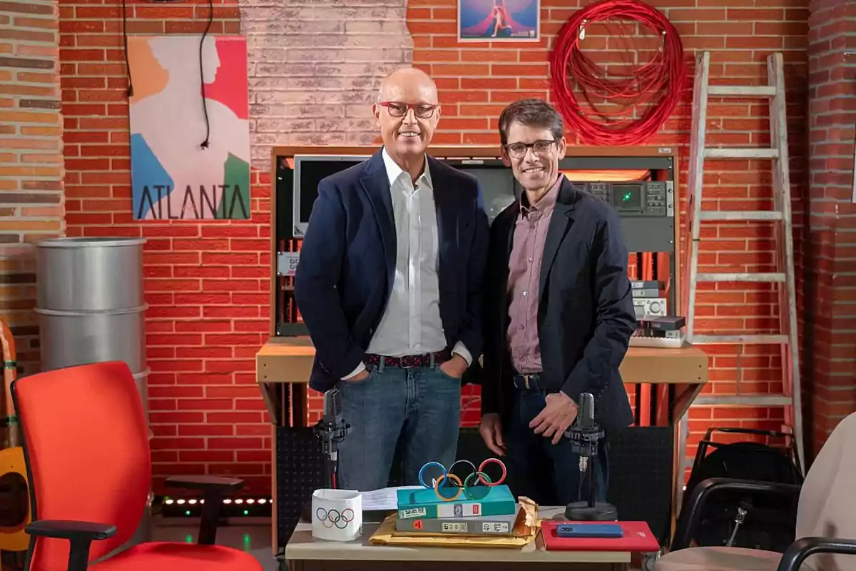 Dos personas posan en un estudio decorado con un fondo de ladrillos, un cartel de Atlanta, una escalera y equipo técnico; en la mesa hay una taza con el logo olímpico, un archivador y micrófonos.