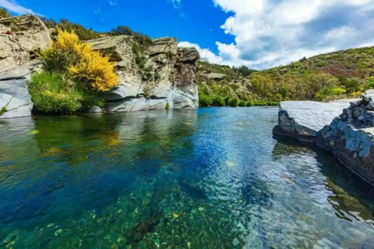 Un río de aguas cristalinas rodeado de rocas y vegetación bajo un cielo azul con nubes.