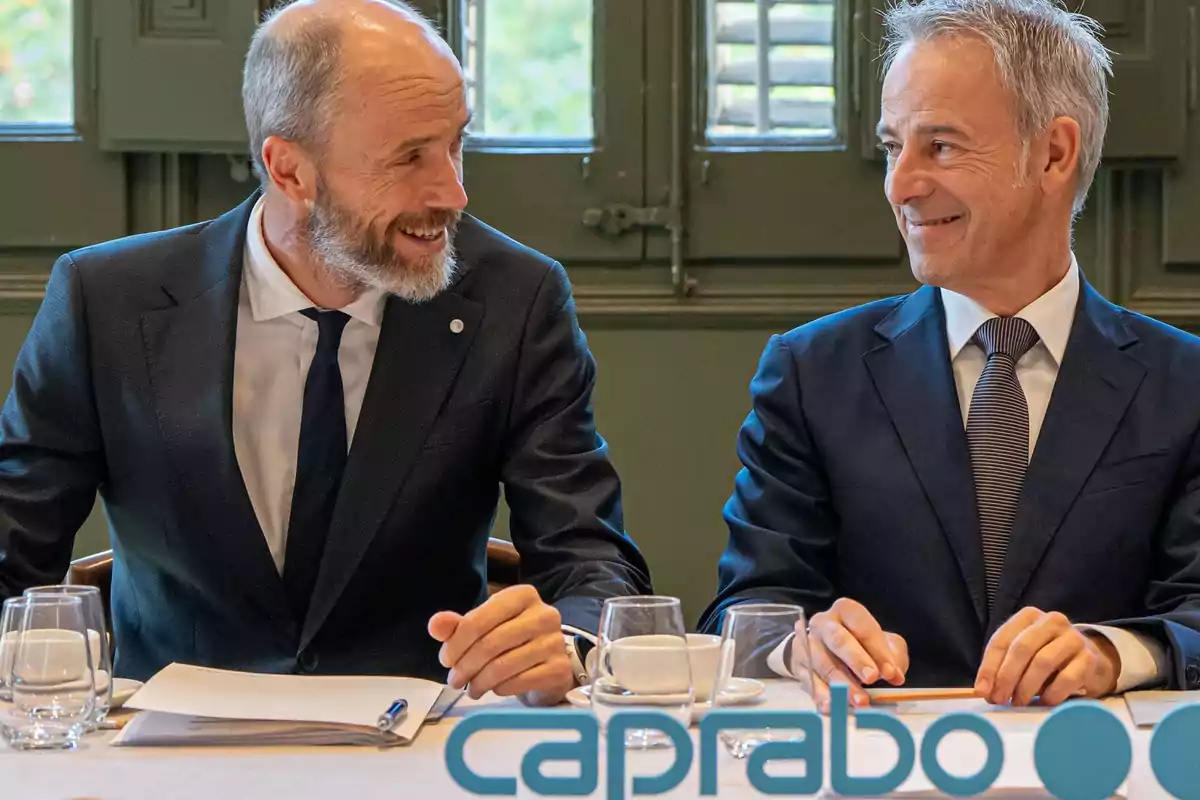 Dos hombres de traje sonríen mientras están sentados en una mesa con documentos y vasos de agua, con el logo de Caprabo en la parte inferior de la imagen.