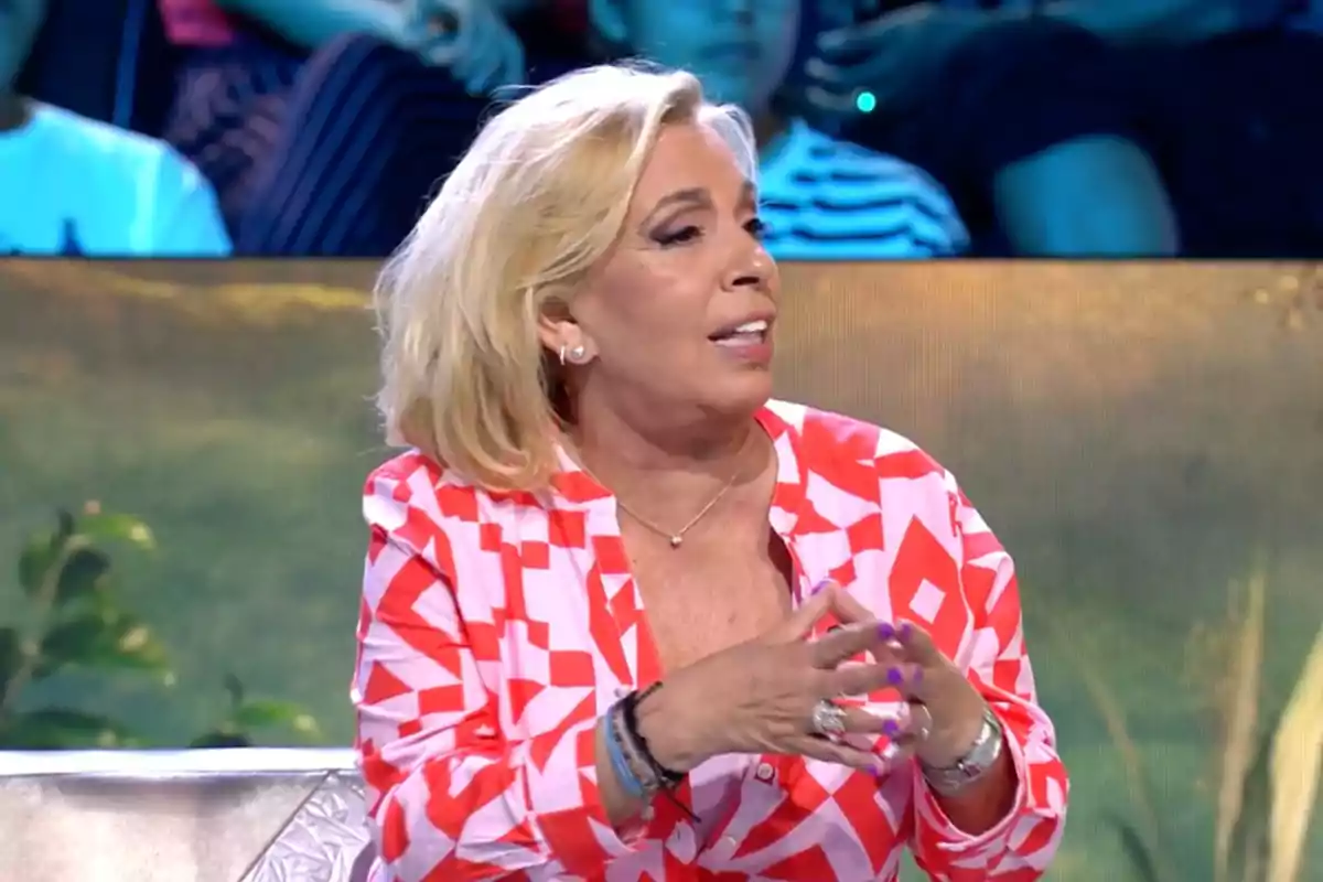 Carmen Borrego con cabello rubio y camisa de patrones geométricos rojos y blancos está hablando en el plató de 'Supervivientes' con un fondo de público desenfocado. Carmen Borrego con cabello rubio y camisa de patrones geométricos rojos y blancos está hablando en el plató de 'Supervivientes' con un fondo de público desenfocado.