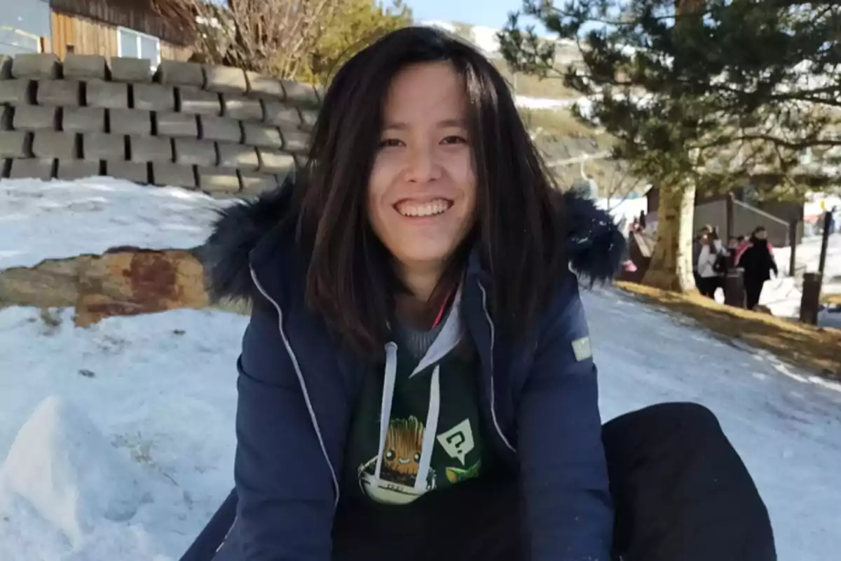 Una persona sonriente con chaqueta azul y sudadera con capucha, sentada en la nieve con un fondo de árboles y una estructura de madera.