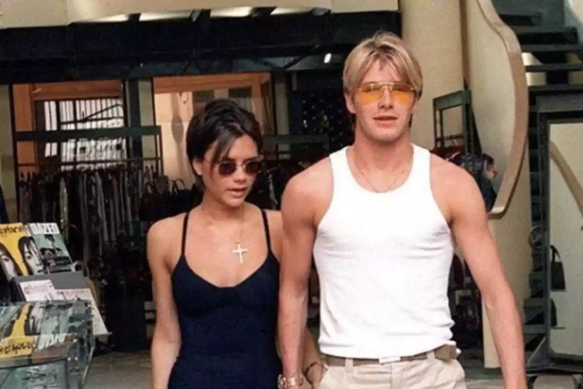 Una pareja caminando juntos, ambos con gafas de sol, ella con un vestido negro y un collar con una cruz, él con una camiseta sin mangas blanca y pantalones claros.