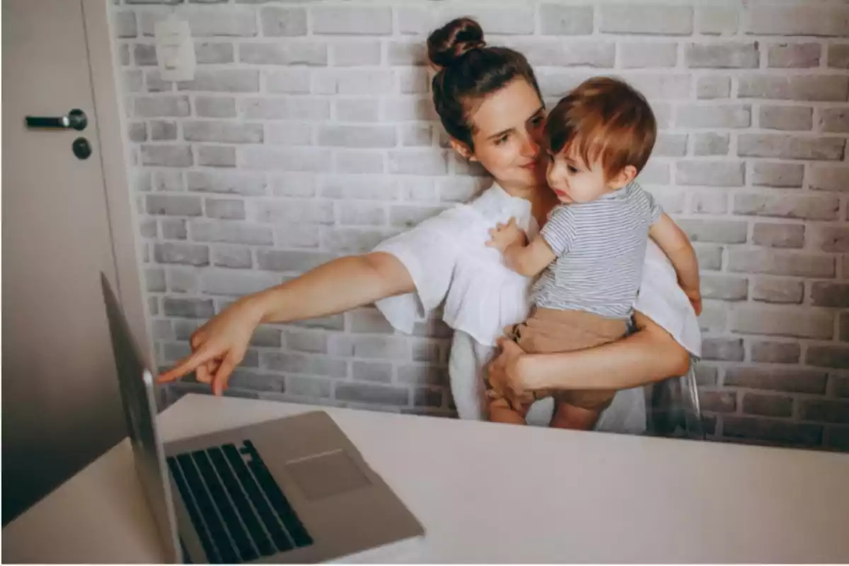 Una mujer sostiene a un niño pequeño mientras señala la pantalla de una computadora portátil en una habitación con una pared de ladrillos blancos.