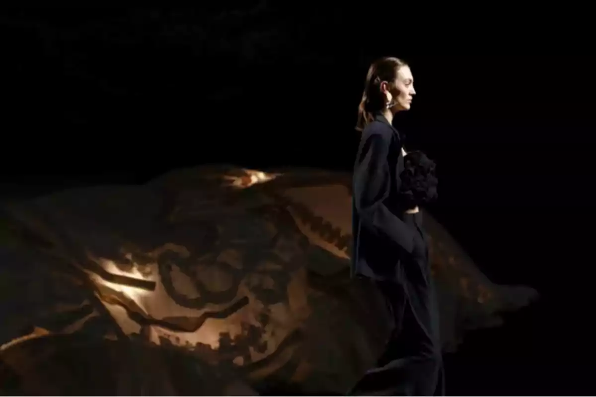 Una modelo camina por la pasarela con un traje oscuro y un fondo artístico iluminado.