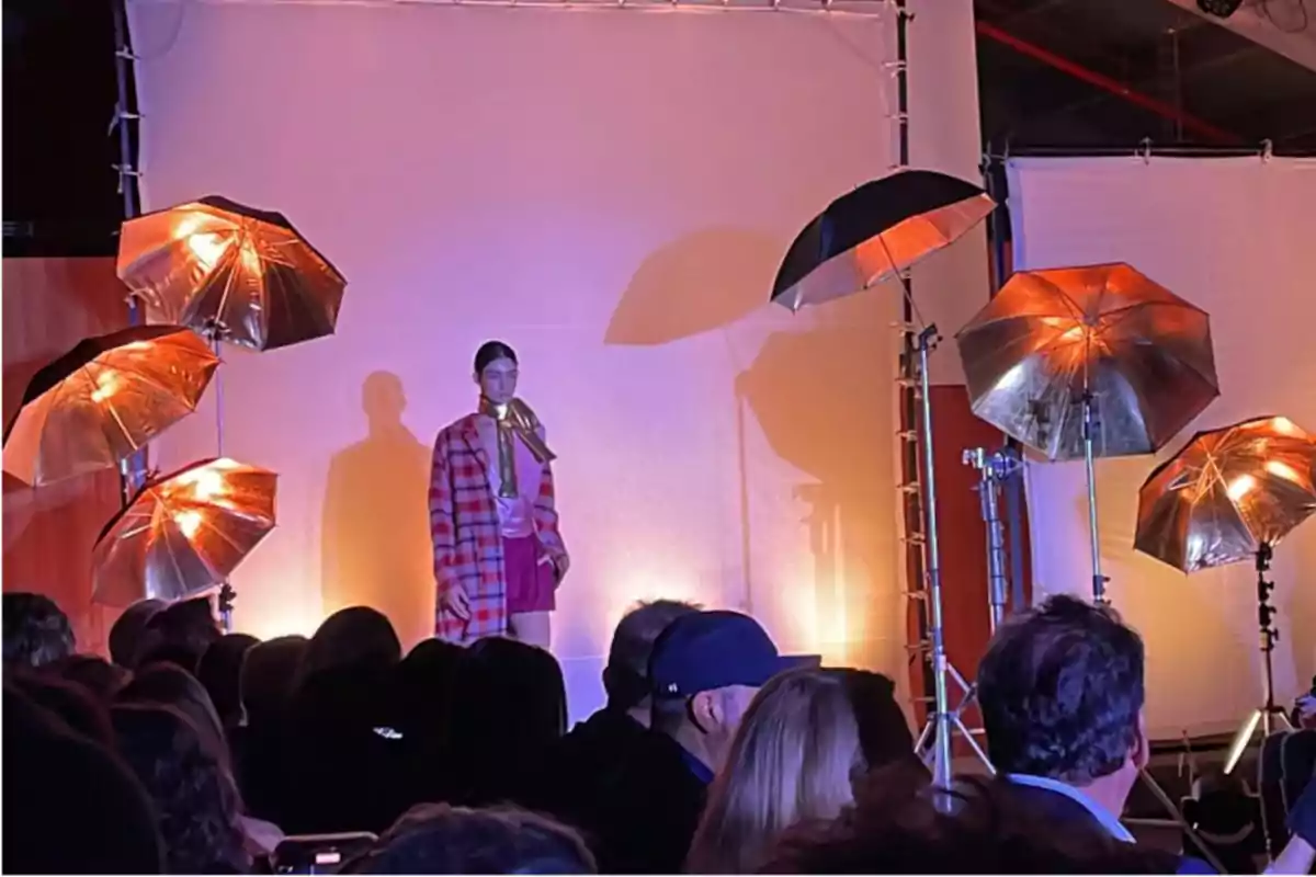 Una modelo en una pasarela de moda iluminada por varios paraguas de luz dorada, con una audiencia observando.