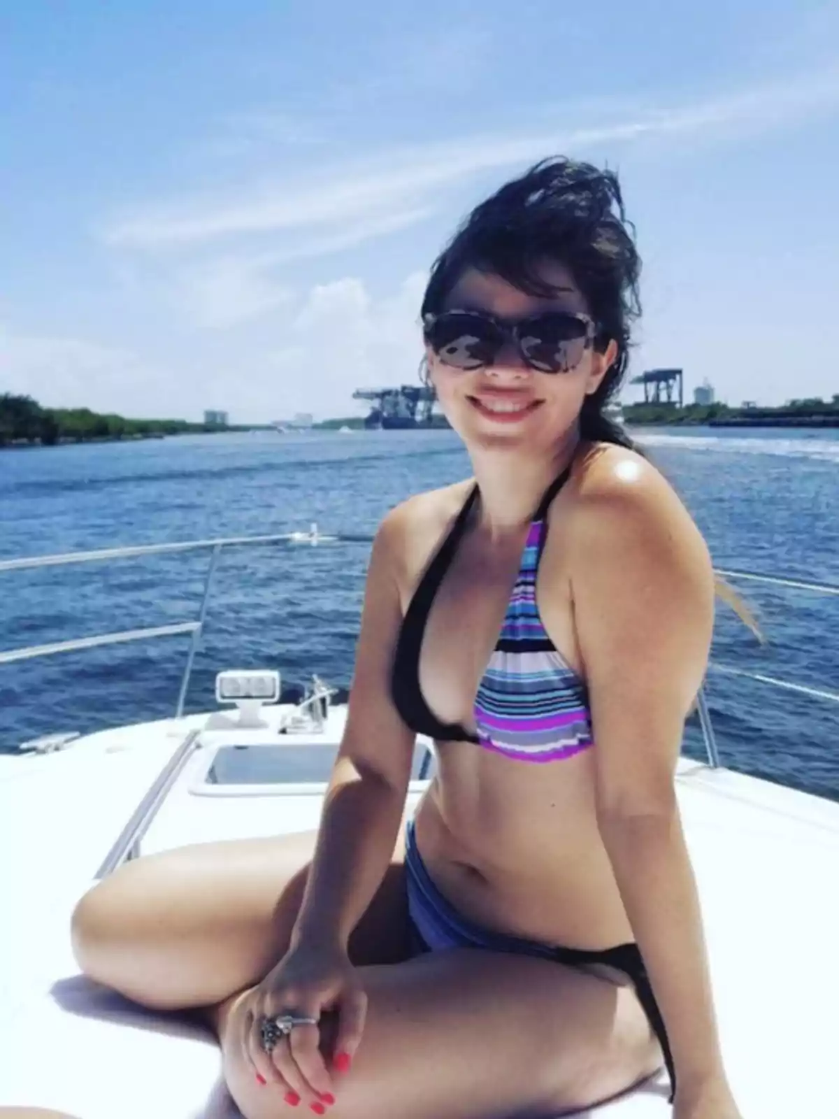 Mujer en bikini con gafas de sol sonriendo mientras está sentada en un bote con el mar y un cielo despejado de fondo.