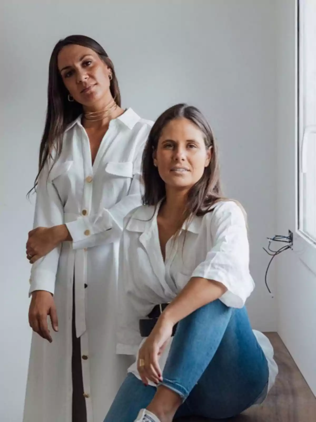 Dos mujeres posando juntas, una de pie y la otra sentada, ambas vestidas con camisas blancas y jeans, en un entorno interior con una pared blanca de fondo.