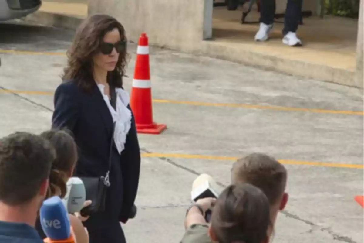 Mujer con gafas de sol y traje oscuro caminando frente a un grupo de periodistas con micrófonos en un área de estacionamiento.