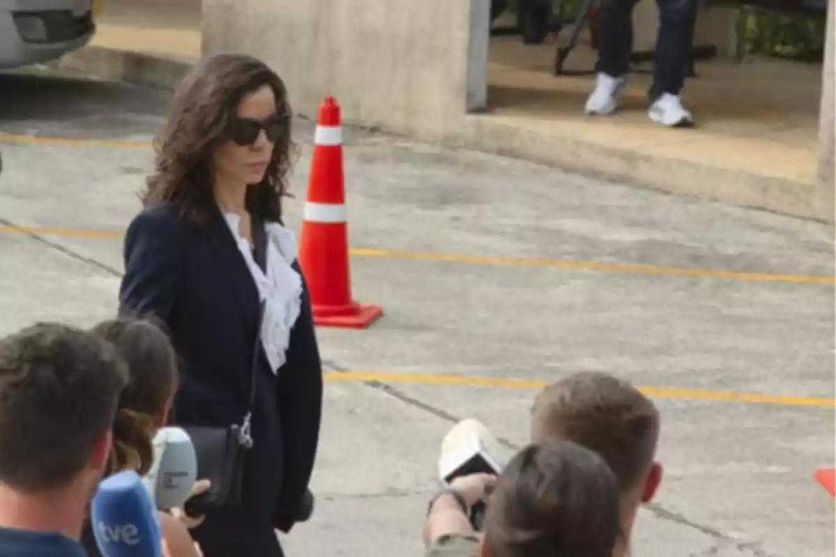 Una mujer con gafas de sol y traje oscuro camina por un estacionamiento mientras es entrevistada por varios periodistas.