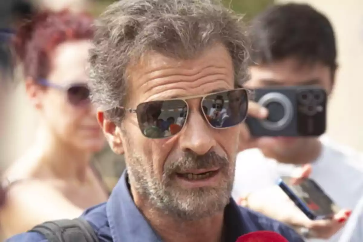 Un hombre con gafas de sol y barba está hablando mientras es fotografiado y grabado por varias personas a su alrededor.